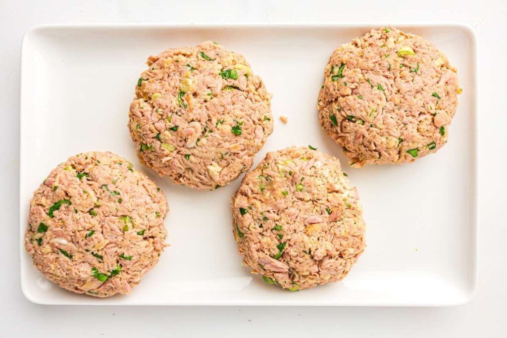 Four uncooked tuna patties mixed with herbs on a rectangular white plate.