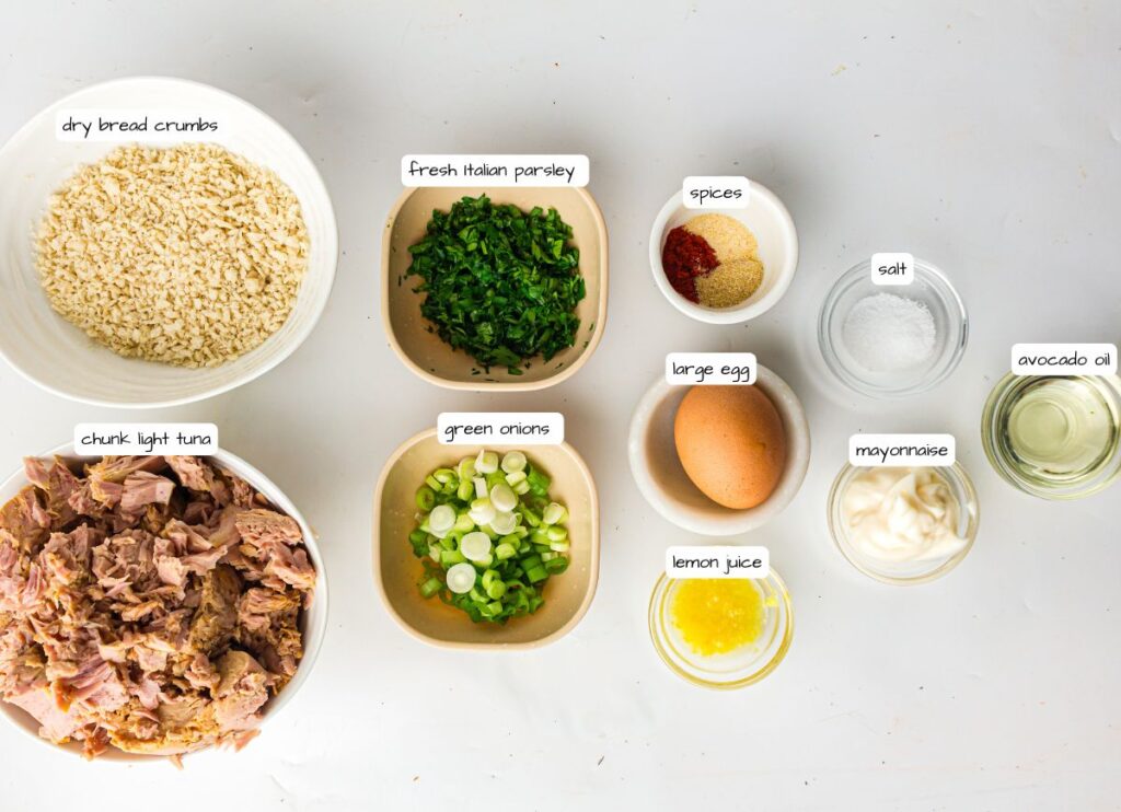 Ingredients for a tuna recipe arranged on a white surface, including bread crumbs, parsley, spices, salt, egg, oil, tuna, green onions, lemon juice, and mayonnaise in separate bowls.