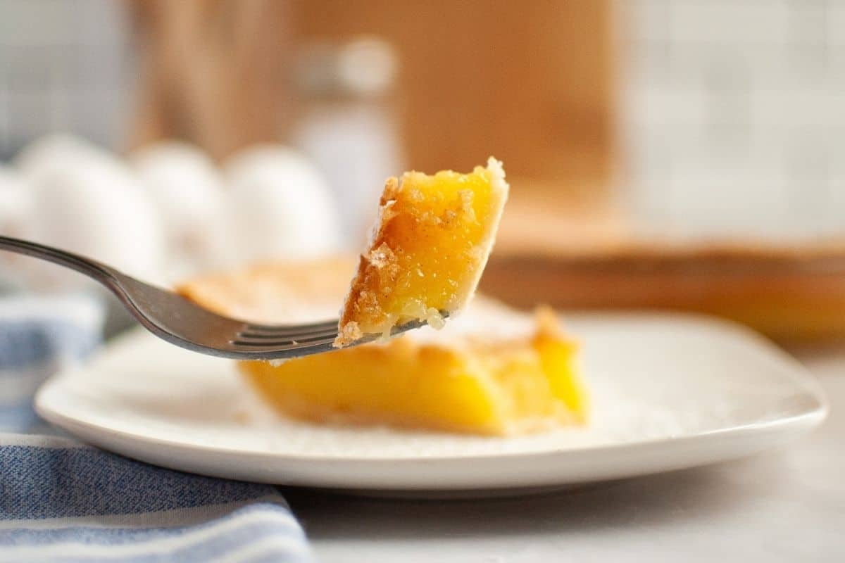 A fork holds a bite of Vinegar pie above a plate with a larger slice, with eggs and kitchen items blurred in the background.