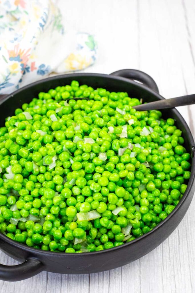 A black bowl filled with cooked green peas and diced onions, with a serving spoon on the side, placed on a white wooden surface.