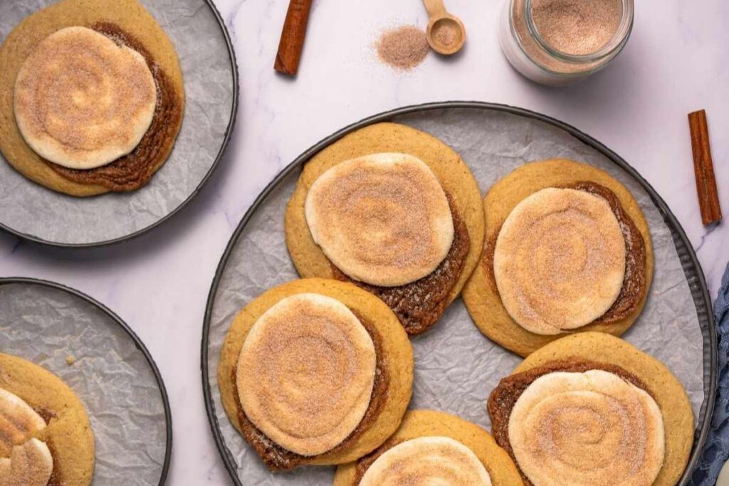 A plate of cinnamon roll cookies topped with swirled icing and sprinkled cinnamon sugar, with extra cinnamon sticks and sugar in the background.