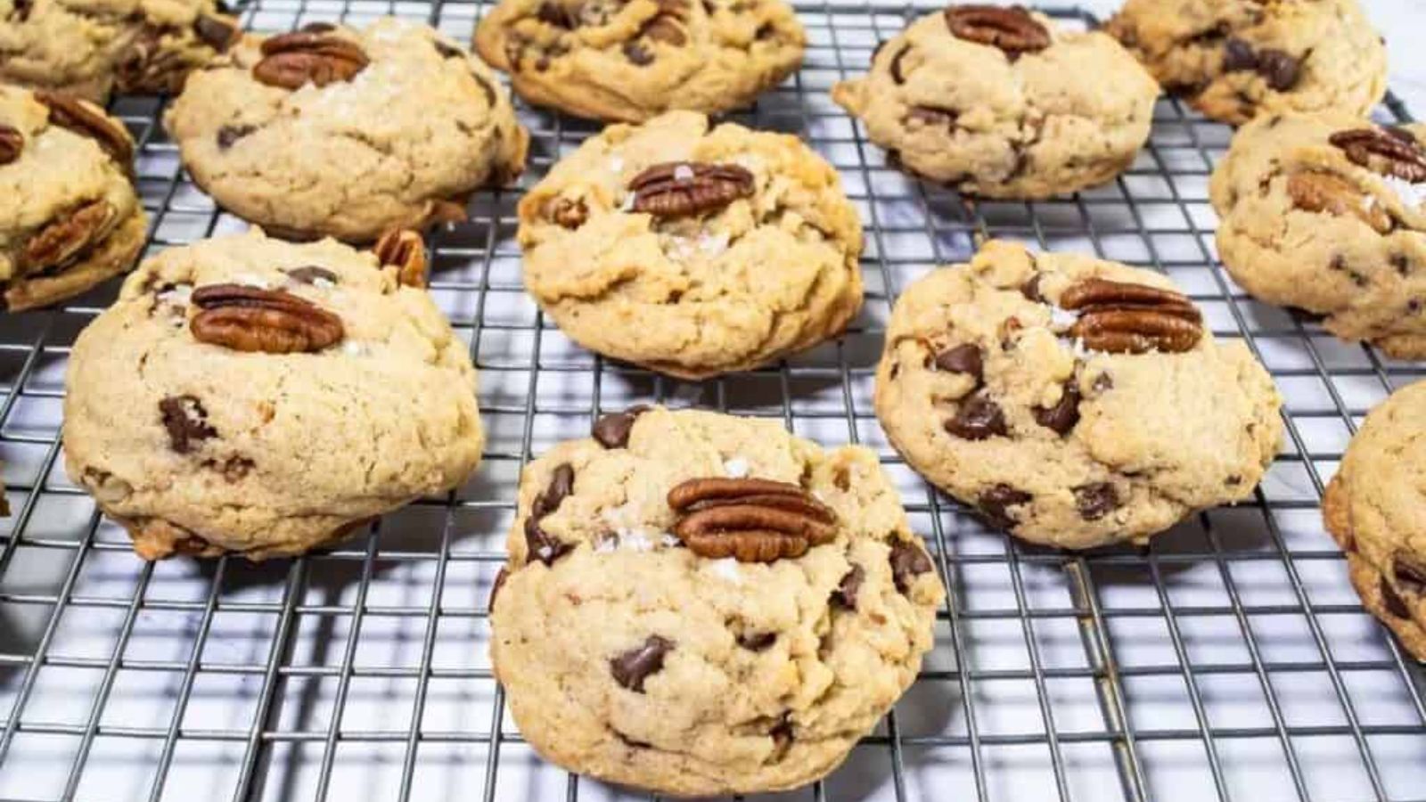 Chocolate chip cookies with pecan halves on top are cooling on a metal wire rack.
