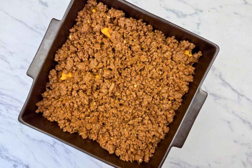 A square baking pan filled with cooked ground beef and small pieces of orange vegetables on a marble countertop.