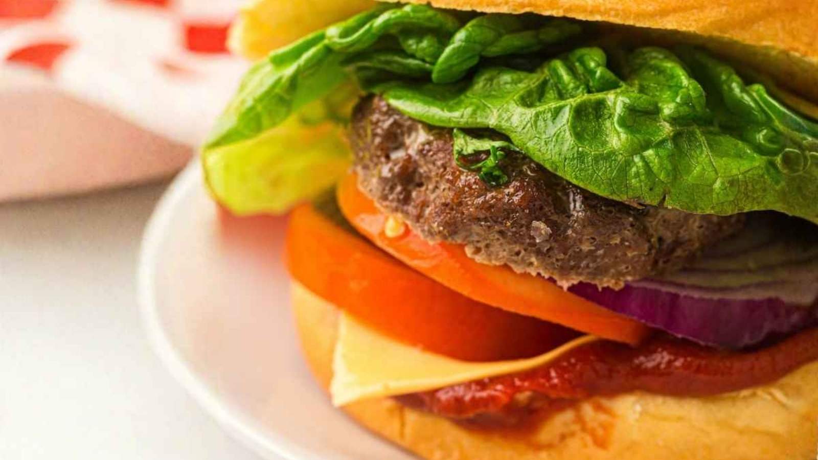 Close-up of a hamburger with lettuce, tomato, onion, cheddar cheese, ketchup, and a beef patty on a bun, served on a white plate.