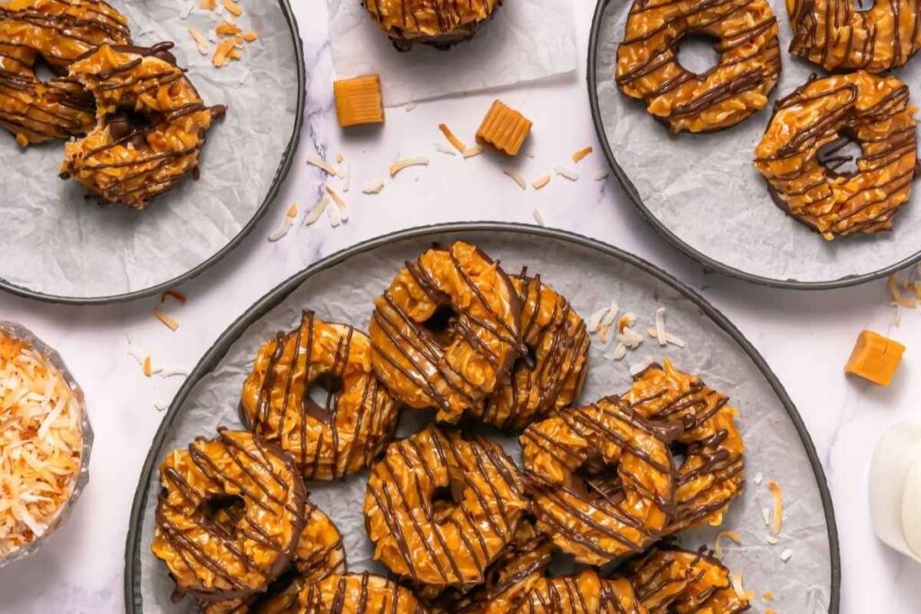 Plates of chocolate-drizzled caramel and coconut ring cookies are arranged on a table, with shredded coconut and caramel candies nearby.