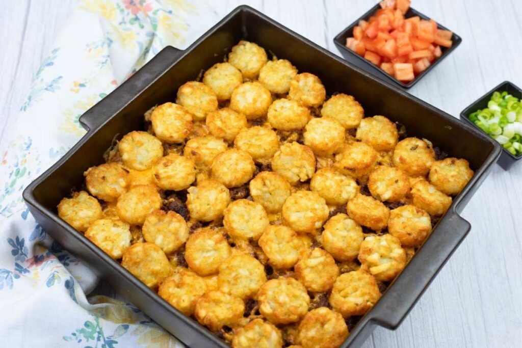 A square baking dish filled with tater tot casserole, topped with golden tater tots, sits on a table next to bowls of diced tomatoes and chopped green onions.