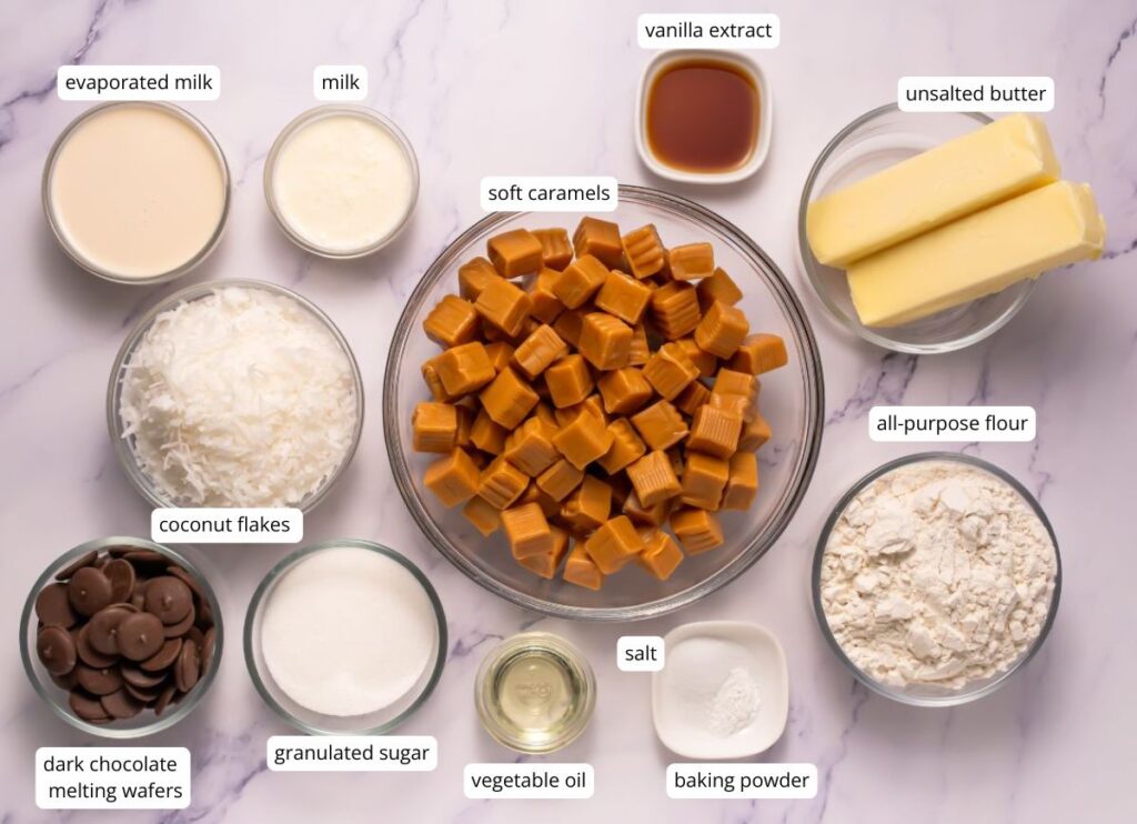 Ingredients for baking are arranged on a marble surface, including soft caramels, flour, butter, coconut flakes, sugar, vanilla extract, baking powder, salt, milk, and chocolate wafers.
