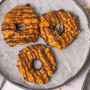 Three round cookies topped with caramel, toasted coconut, and drizzled with chocolate are arranged on a parchment-lined plate.
