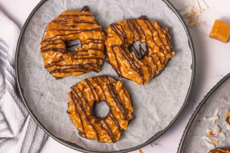 Three round cookies topped with caramel, toasted coconut, and drizzled with chocolate are arranged on a parchment-lined plate.