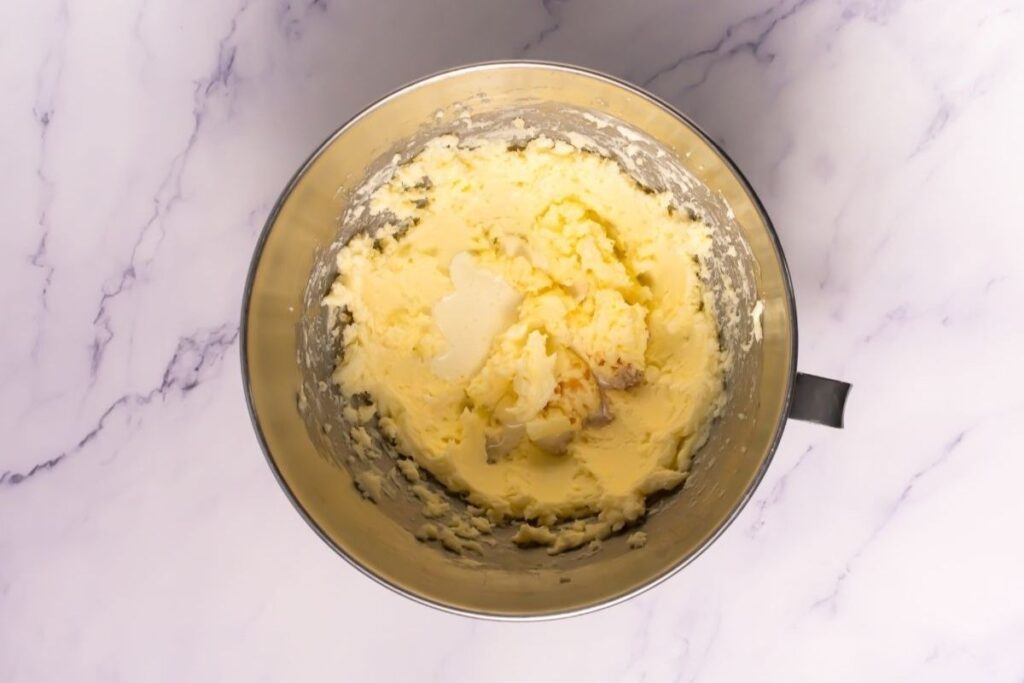 A metal mixing bowl containing creamed butter and sugar sits on a white marble surface.