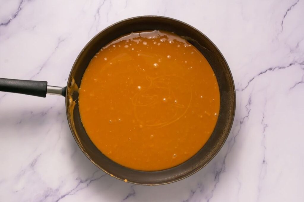 A nonstick frying pan filled with smooth, orange-brown sauce sits on a white marble surface.
