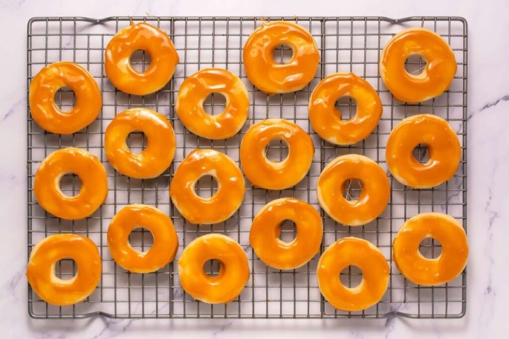 Eighteen glazed donuts arranged in rows on a cooling rack over a marble surface.