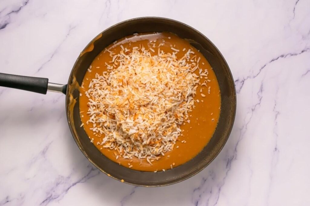 A skillet filled with caramel sauce topped with a pile of shredded toasted coconut, sitting on a white marble surface.