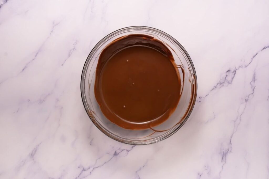 A glass bowl filled with melted chocolate sits on a white marble surface.