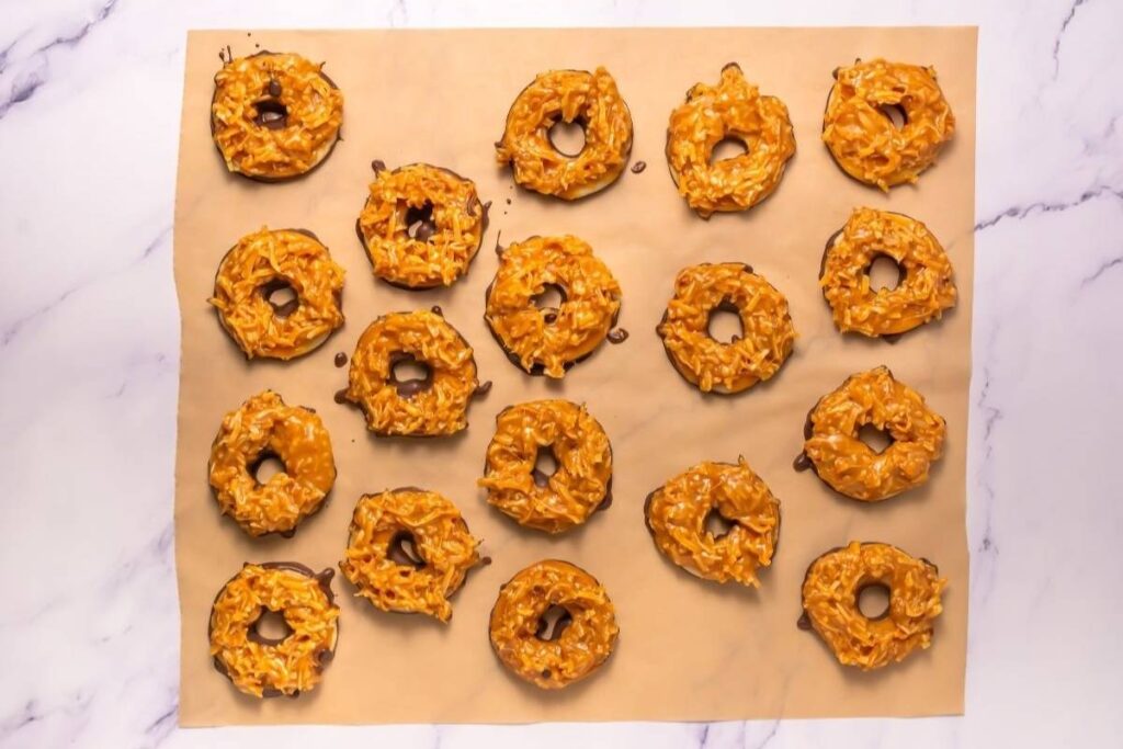 Eighteen caramel and coconut-covered ring cookies, some with chocolate bases, are arranged on a sheet of parchment paper on a marble surface.