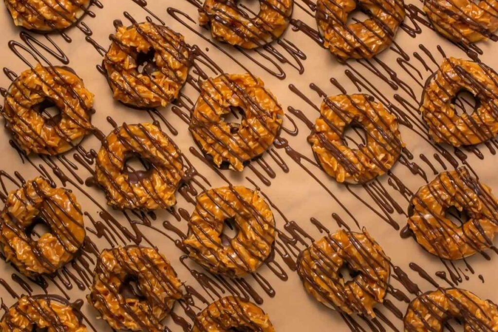 Rows of caramel and coconut-covered ring cookies are drizzled with chocolate on a tan surface.