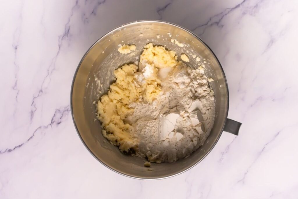 A metal mixing bowl containing partially mixed cookie dough, with flour and creamed butter and sugar visible, on a white marble surface.