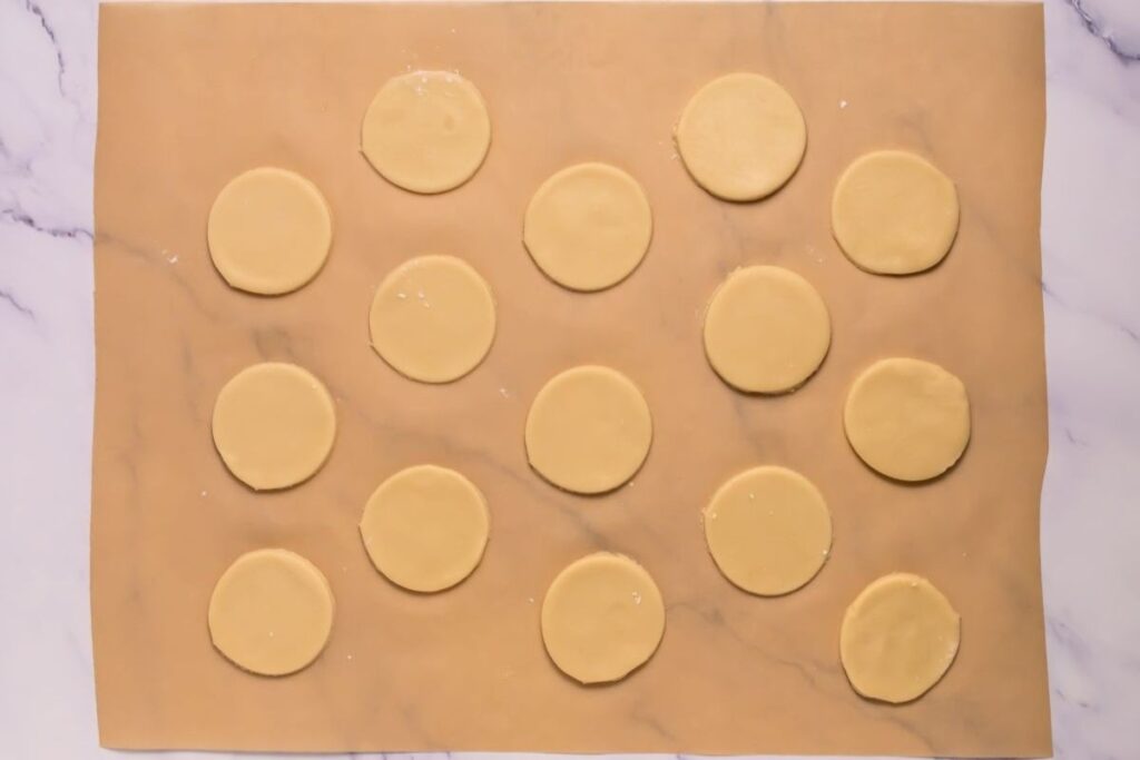 Fifteen round cookie dough pieces are evenly spaced on a sheet of rolled dough on a marble surface.