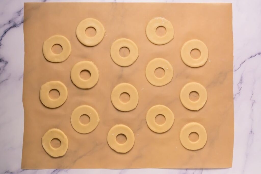 Round dough shapes with holes in the center are arranged on a sheet of parchment paper on a marble surface.