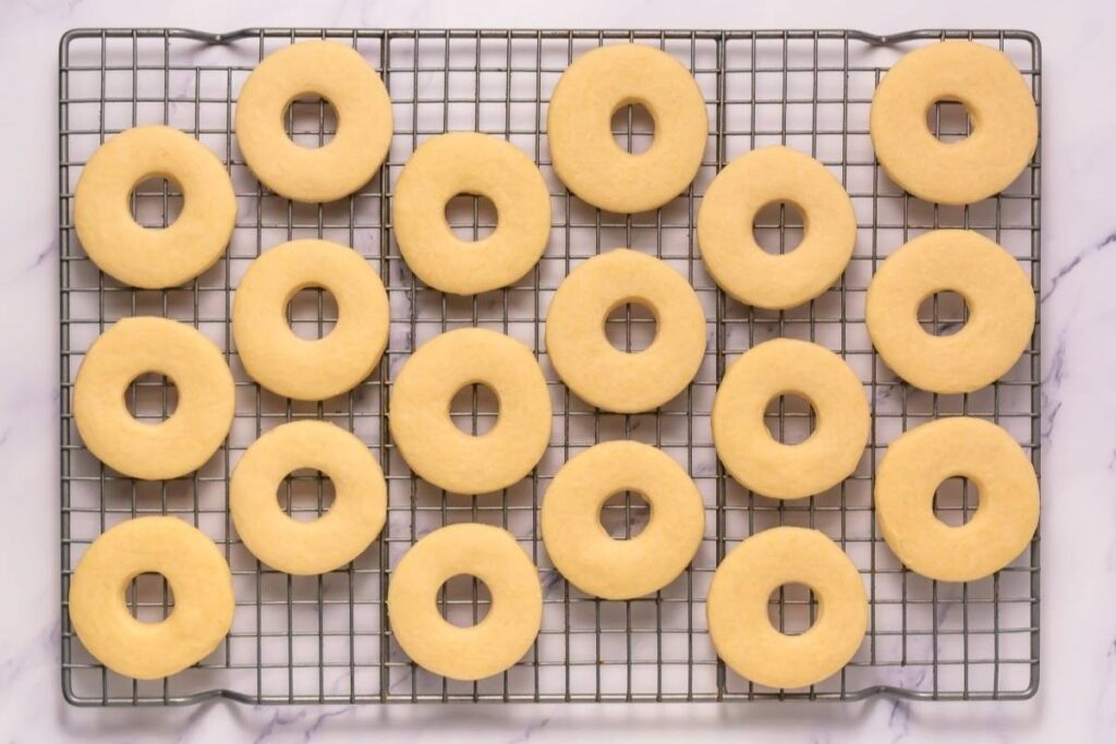Round cookies with holes in the center are arranged in neat rows on a wire cooling rack over a white surface.