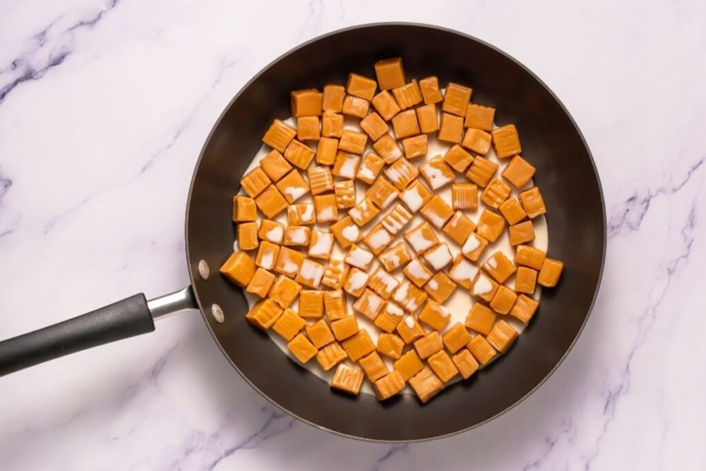 A frying pan filled with cubed caramel candies and a layer of milk on a white marble surface.