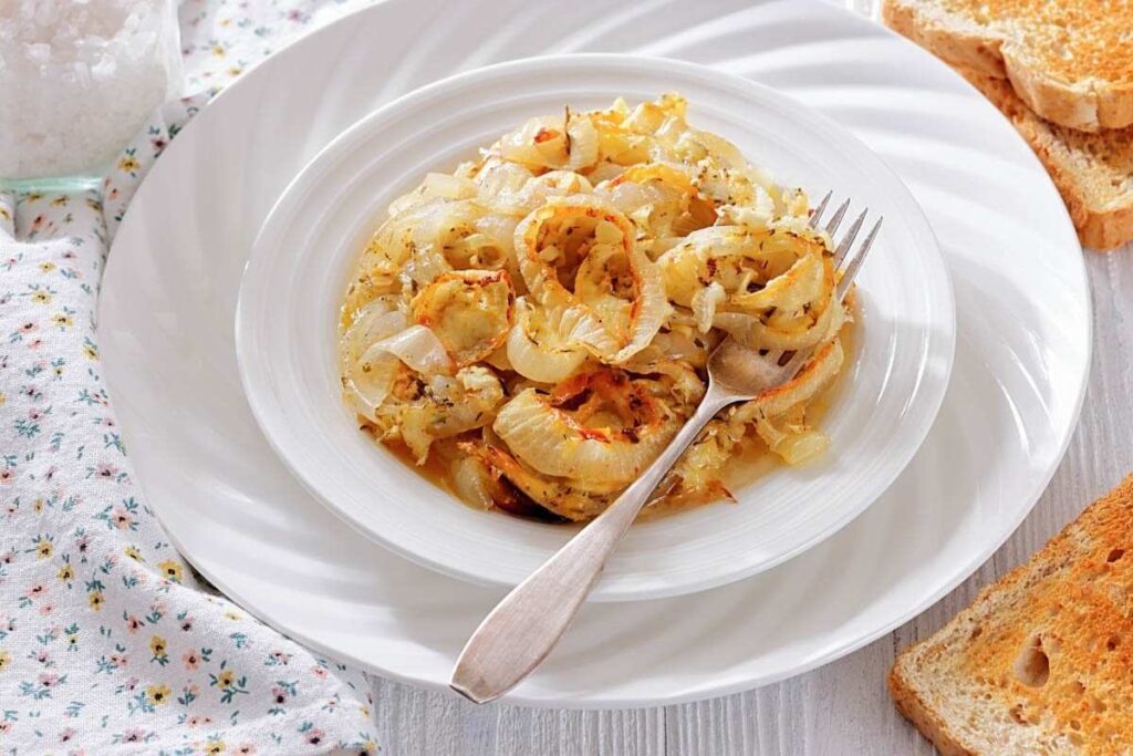 A white plate with cooked sliced onions, seasoned with herbs, sits on a table next to toasted bread and a floral napkin. A fork rests on the plate.