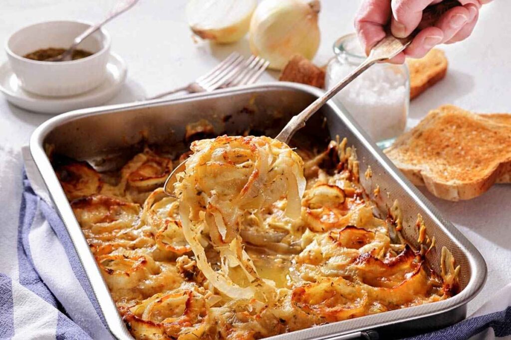 A hand serves a portion of baked scalloped potatoes with onions and cheese from a rectangular baking dish, surrounded by toast, fork, and seasoning.