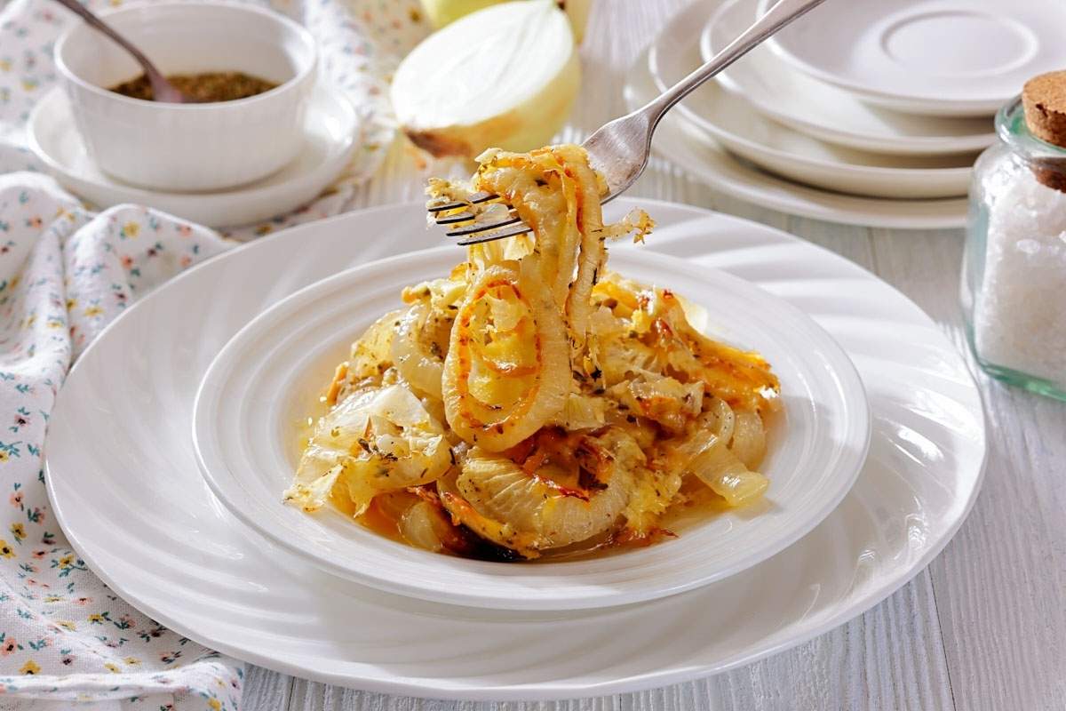 A plate of baked fish with lemon slices and onions is being lifted by a fork, with plates, a salt jar, and a bowl in the background.