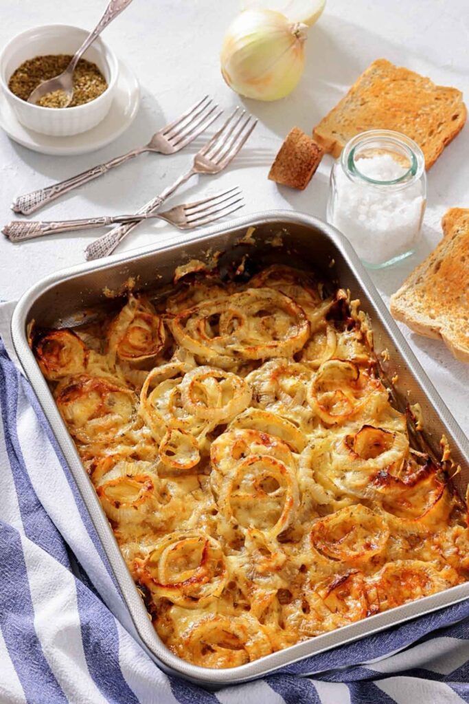A baked casserole topped with browned onion rings in a metal pan, surrounded by toast, salt, a whole onion, spice bowl, and several forks on a striped kitchen towel.
