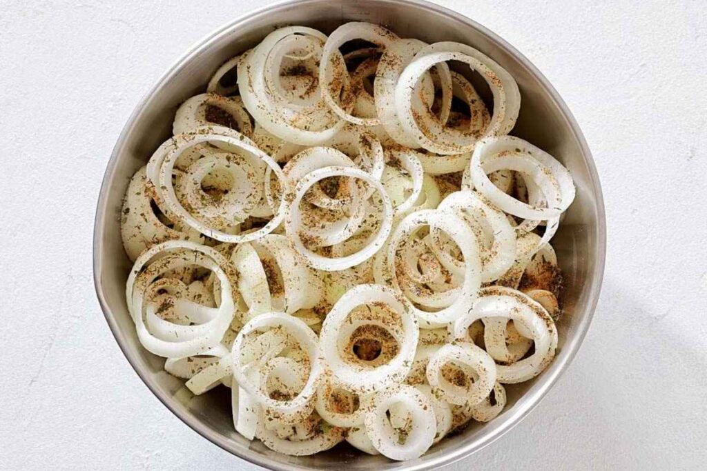 A metal bowl filled with raw onion rings sprinkled with black pepper and spices, placed on a white surface.