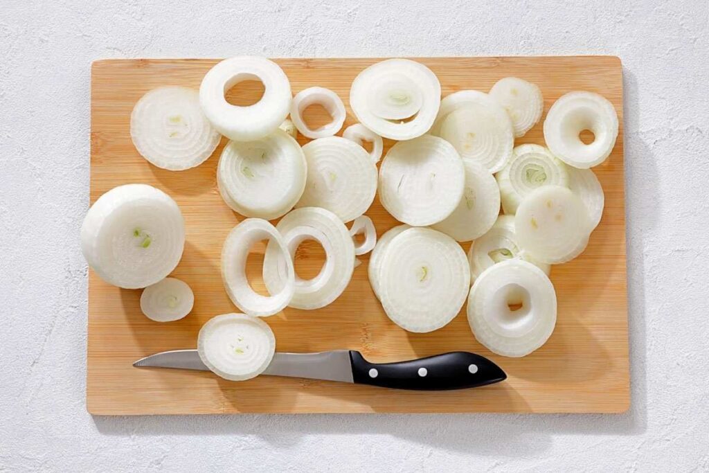 Sliced white onions and a paring knife on a wooden cutting board.