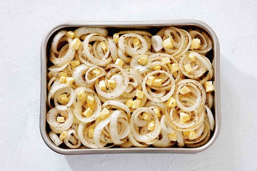 Sliced onions topped with cubes of butter and sprinkled with herbs in a rectangular baking dish on a light surface.