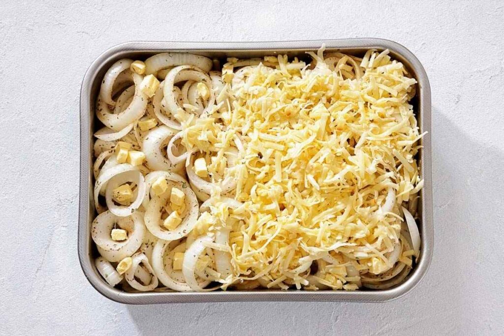 A baking dish filled with sliced onions, butter cubes, and shredded cheese, with half the onions covered in cheese and seasonings visible.