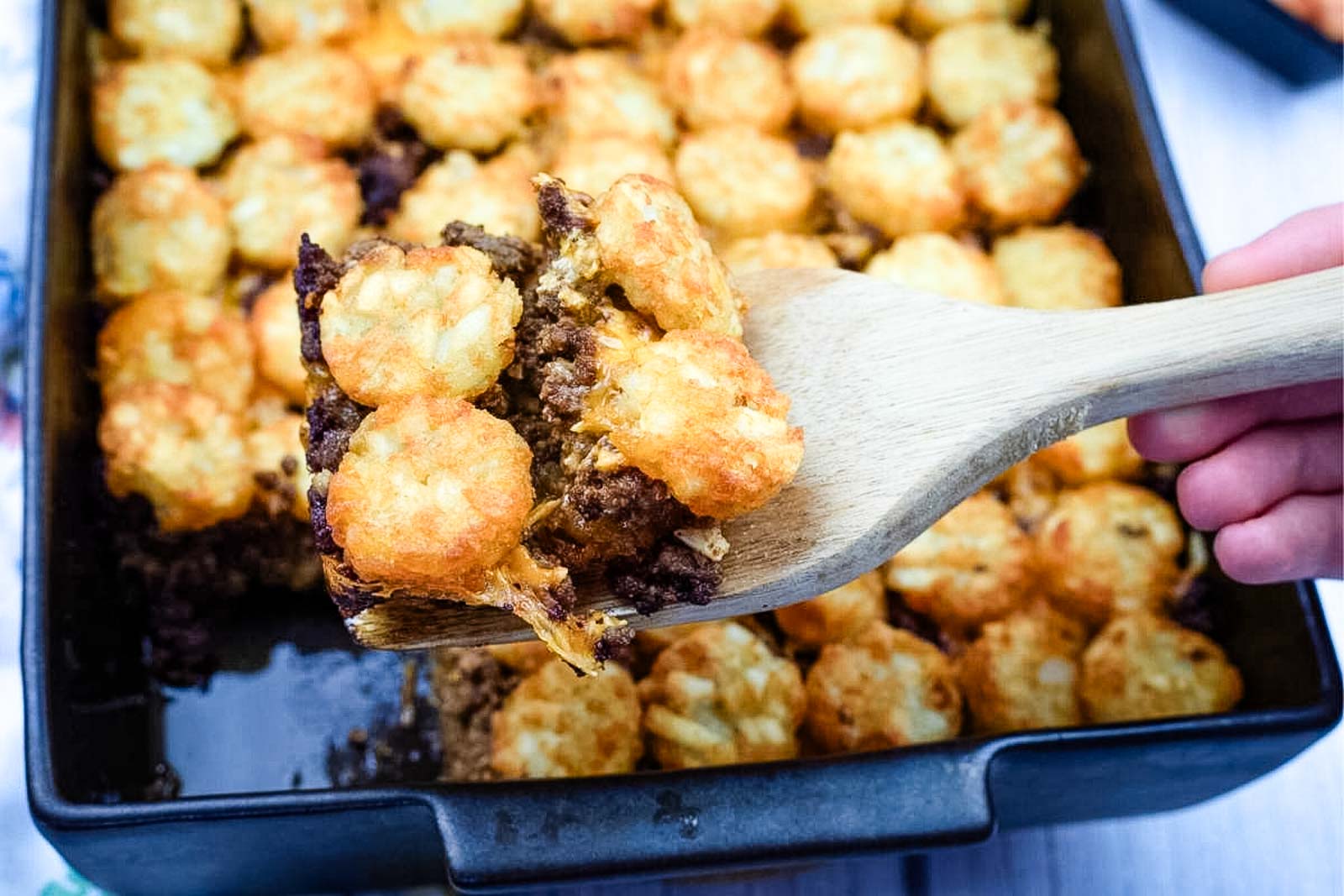 A wooden spatula lifts a serving of tater tot casserole with browned beef and melted cheese from a baking dish.