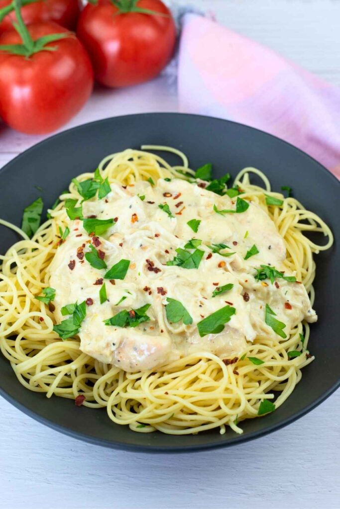 A black bowl of spaghetti topped with creamy chicken, garnished with chopped parsley and red pepper flakes, with tomatoes and a pink cloth in the background.