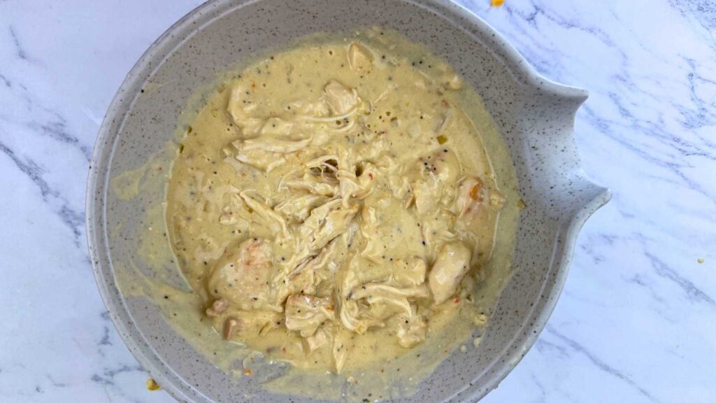 A gray bowl filled with shredded chicken mixed in a creamy yellow sauce sits on a white marble surface.