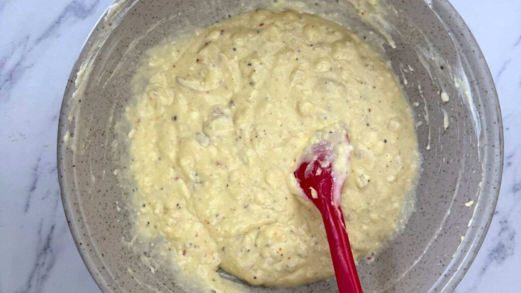 A mixing bowl containing thick, lumpy batter is being stirred with a red spatula on a white marble surface.