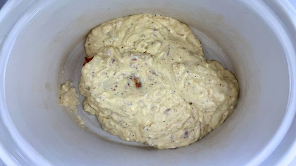 A thick, beige batter with visible seasoning and small chunks sits in the bottom of a white slow cooker.
