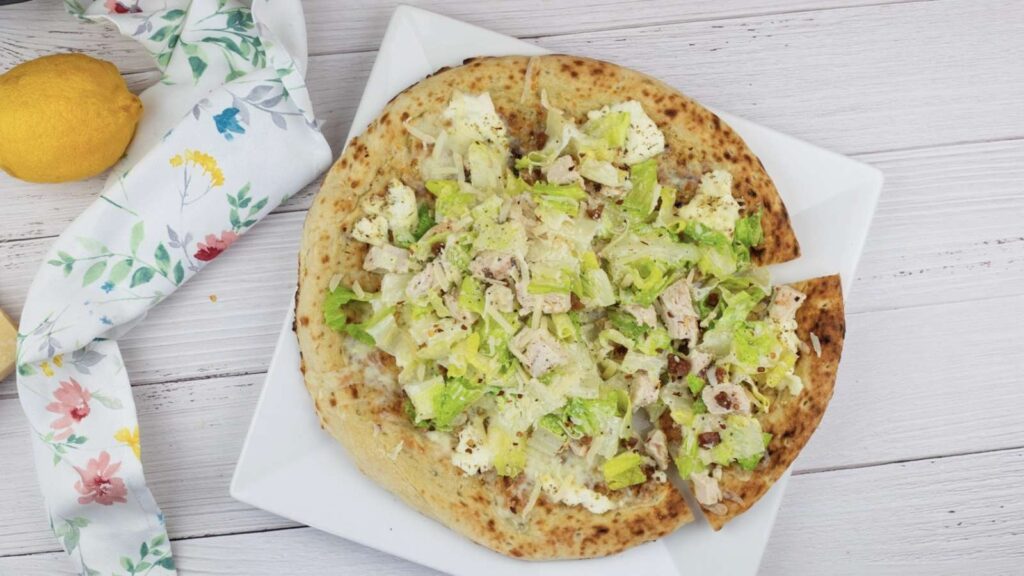 Square white plate with a flatbread topped with lettuce, chicken pieces, cheese, and dressing. A lemon and a floral napkin are nearby on a white wooden surface.
