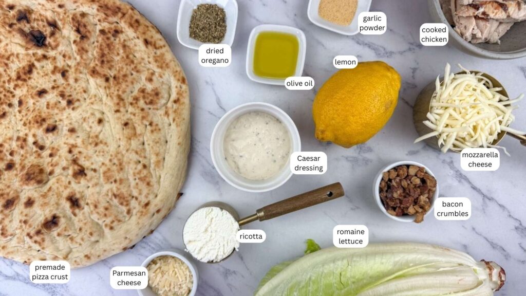 Ingredients for a chicken Caesar pizza arranged on a marble counter, including premade crust, cheeses, bacon, chicken, lettuce, lemon, olive oil, seasonings, and Caesar dressing.