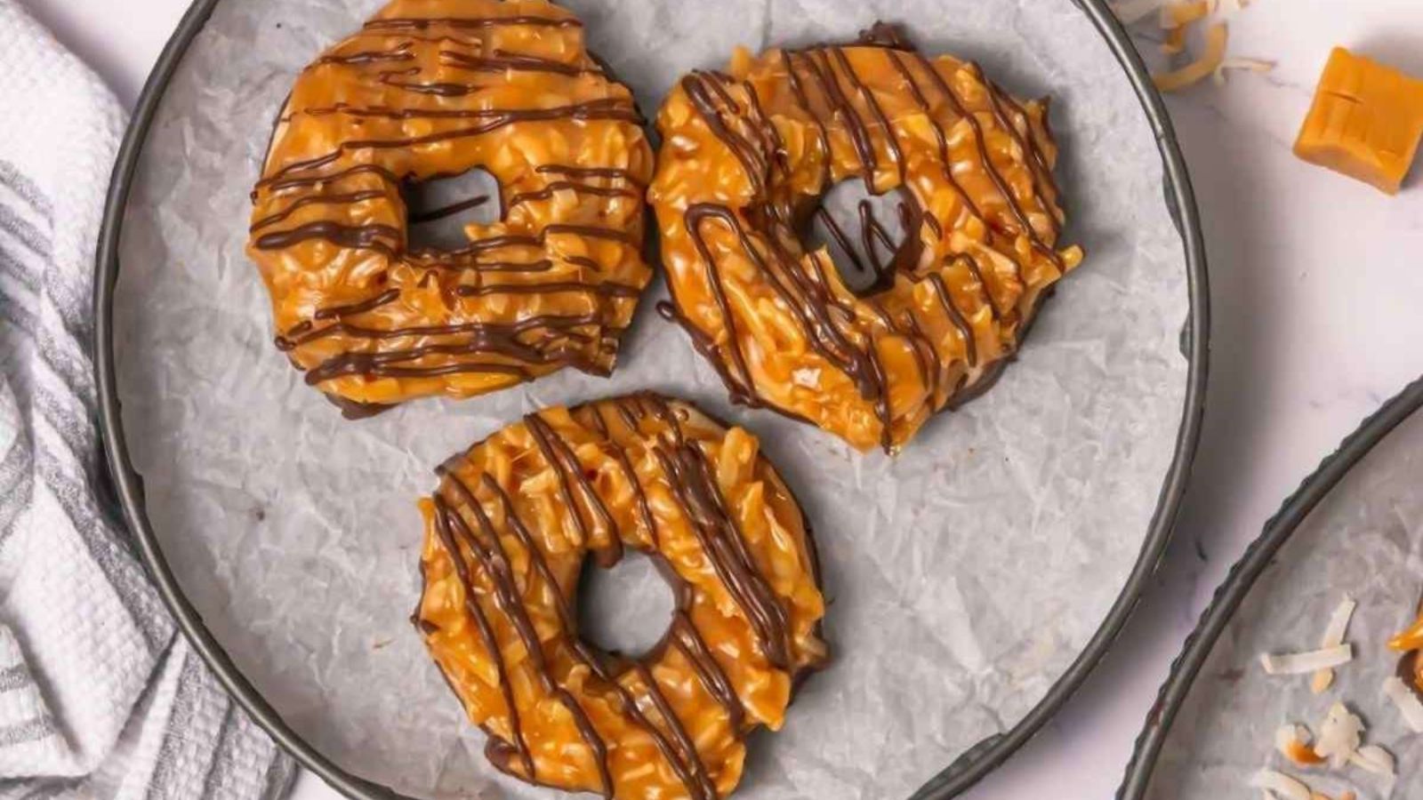 Three caramel and chocolate-drizzled cookies with coconut topping are arranged on a parchment-lined plate.