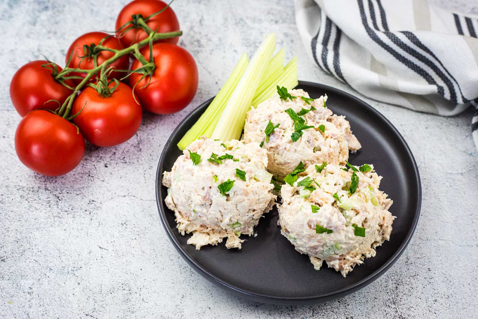 A black plate with three scoops of Copycat Costco Chicken Salad garnished with parsley, celery sticks, and a bunch of tomatoes on the side. A striped towel is in the background.