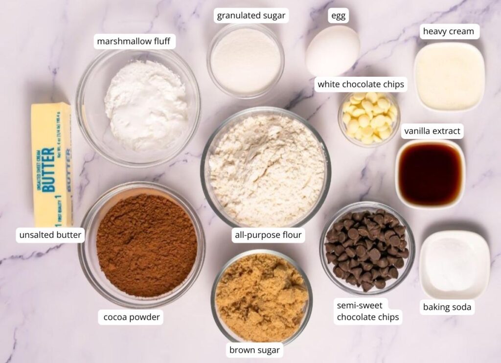 Ingredients for baking cookies arranged in bowls on a marble surface, including butter, sugars, flour, cocoa powder, chocolate chips, marshmallow fluff, egg, cream, vanilla, and baking soda.