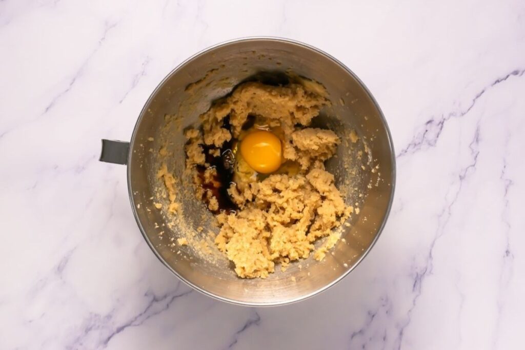 A metal mixing bowl containing cookie dough with an egg and vanilla extract on top, placed on a white marble surface.