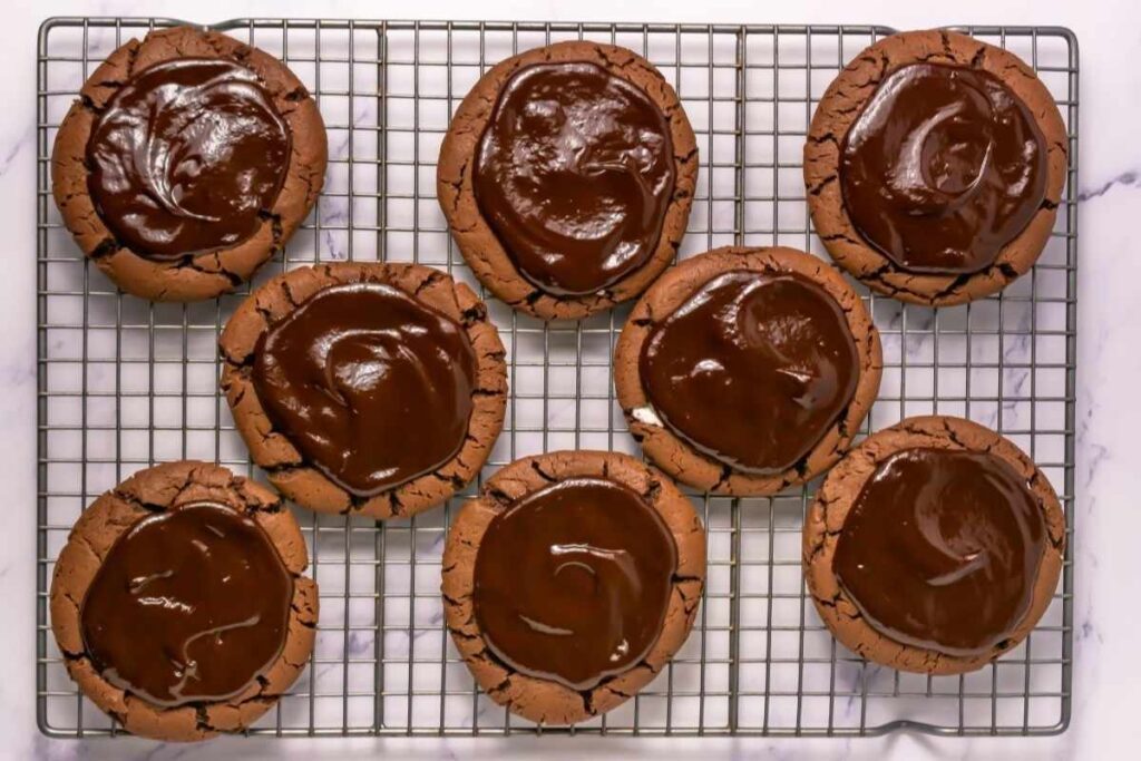 Eight chocolate cookies with a glossy chocolate glaze are arranged on a wire cooling rack over a marble surface.
