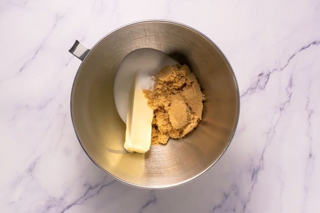 A metal mixing bowl with a stick of butter, white sugar, and brown sugar on a marble surface.