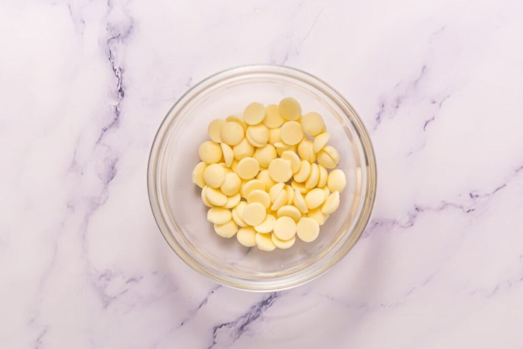 A clear glass bowl filled with white chocolate chips sits on a white marble surface.