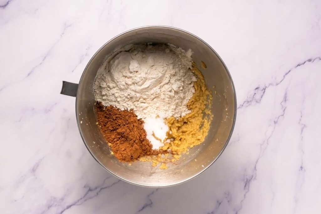 A metal mixing bowl containing flour, cocoa powder, baking powder, and creamed butter and sugar on a marble countertop.