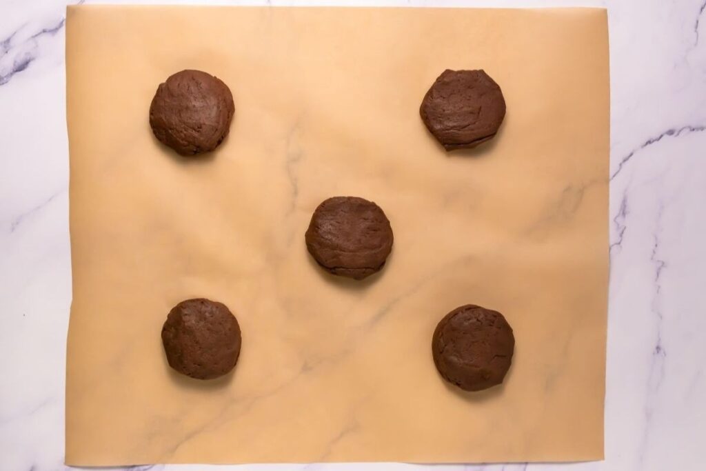 Five evenly spaced chocolate cookie dough balls are placed on a sheet of parchment paper on a marble surface.