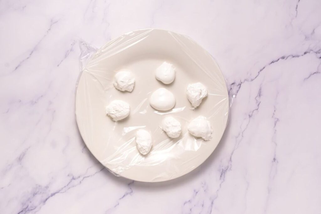 A white plate with seven balls of mozzarella cheese covered in plastic wrap, placed on a light marble surface.